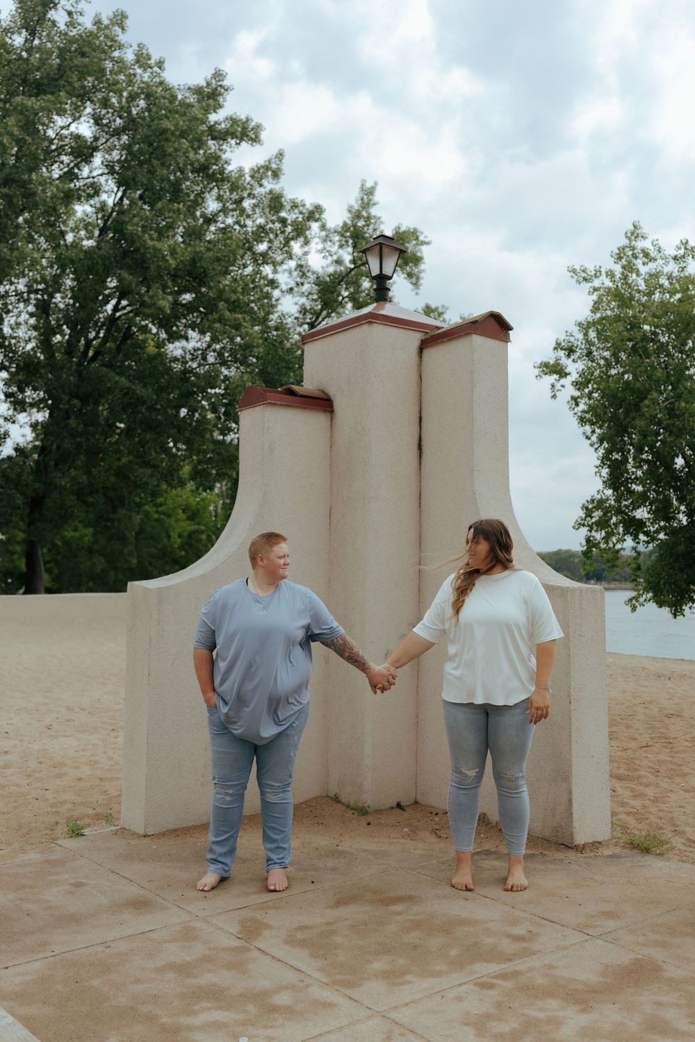 Two people in casual attire stand together against white walls in a desert-like setting with trees in the background.