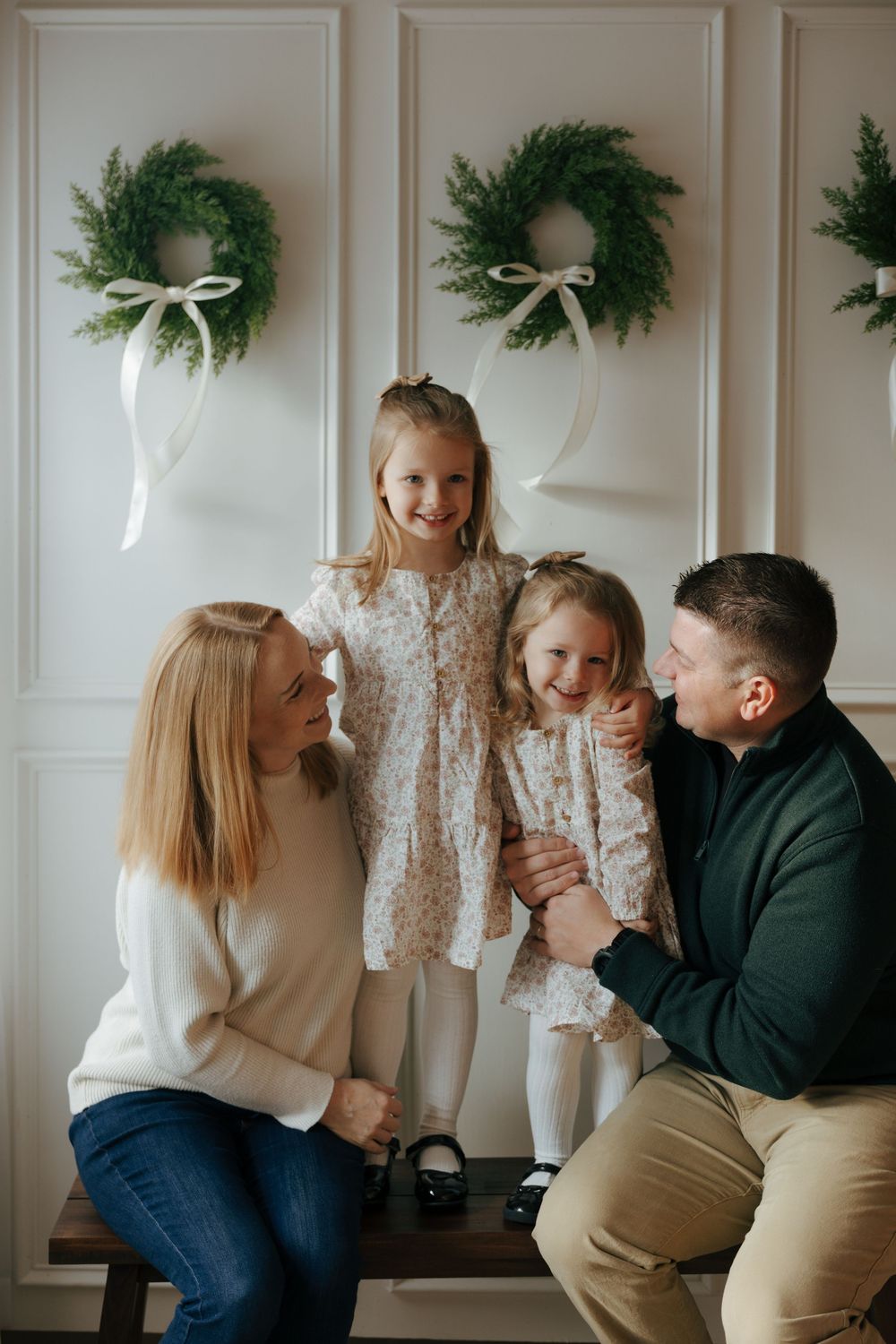 A family sits together on a white bench against a wall decorated with holiday wreaths and white bows.
