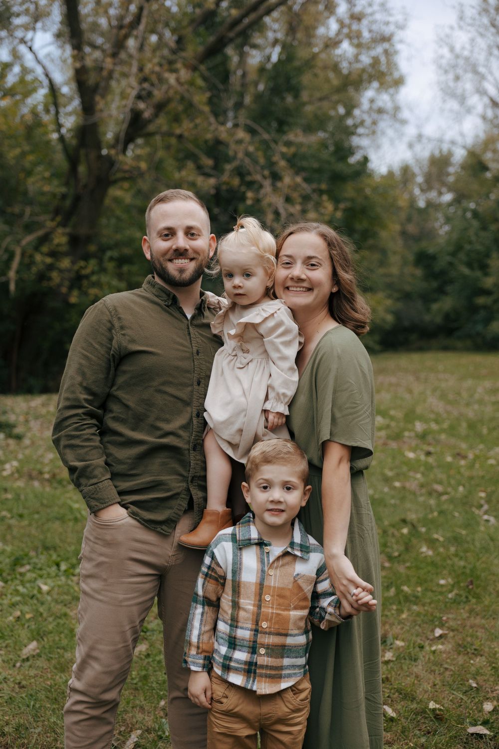 A family of four poses together outdoors on an autumn day with fallen leaves and trees in the background.