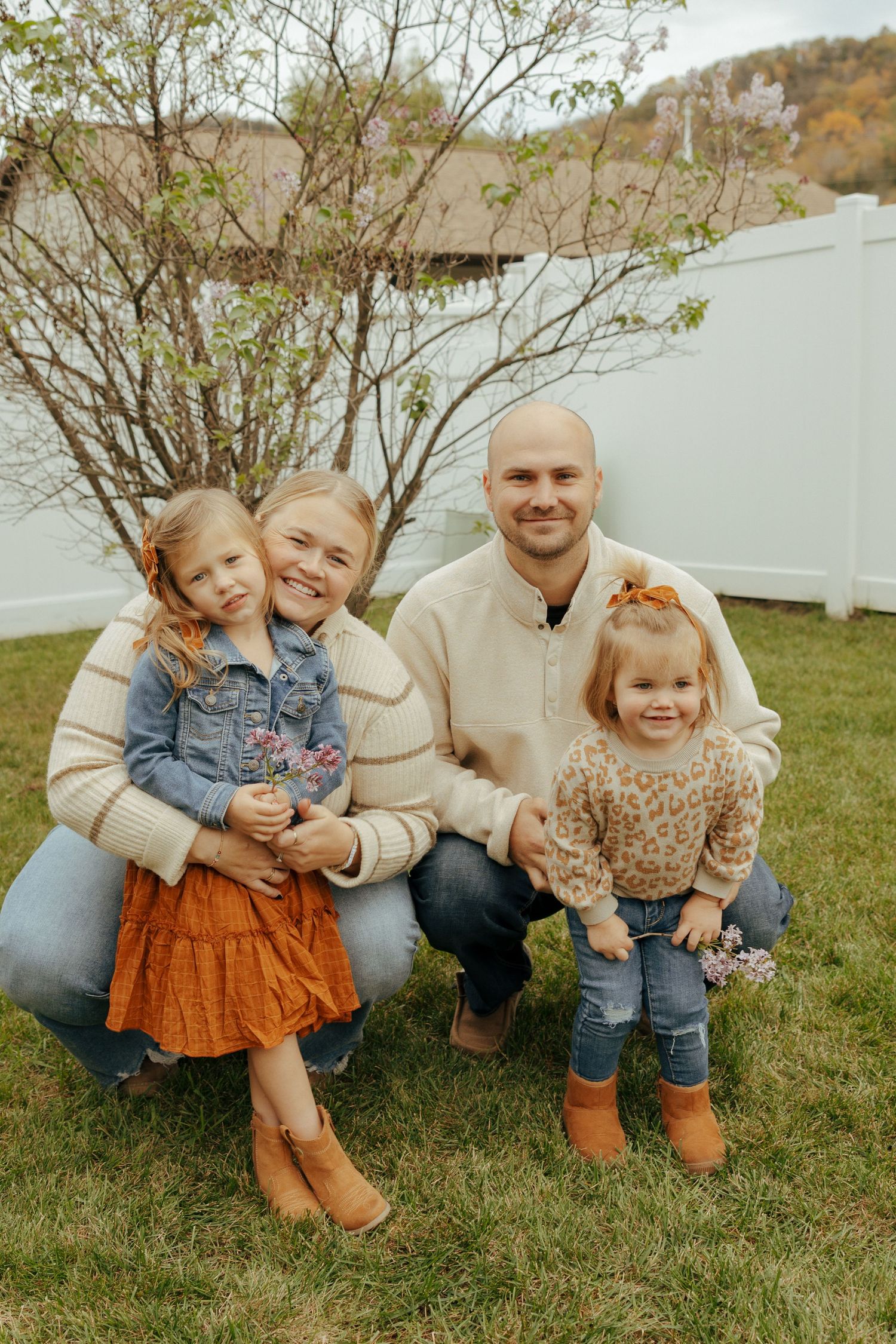 A family poses together on grass in front of a white fence during autumn, wearing coordinated casual fall outfits.
