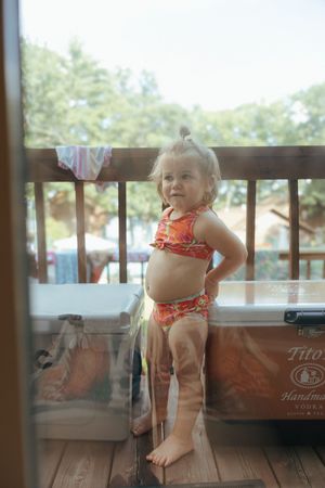 A toddler in an orange swimsuit plays on a wooden deck during a sunny summer day.