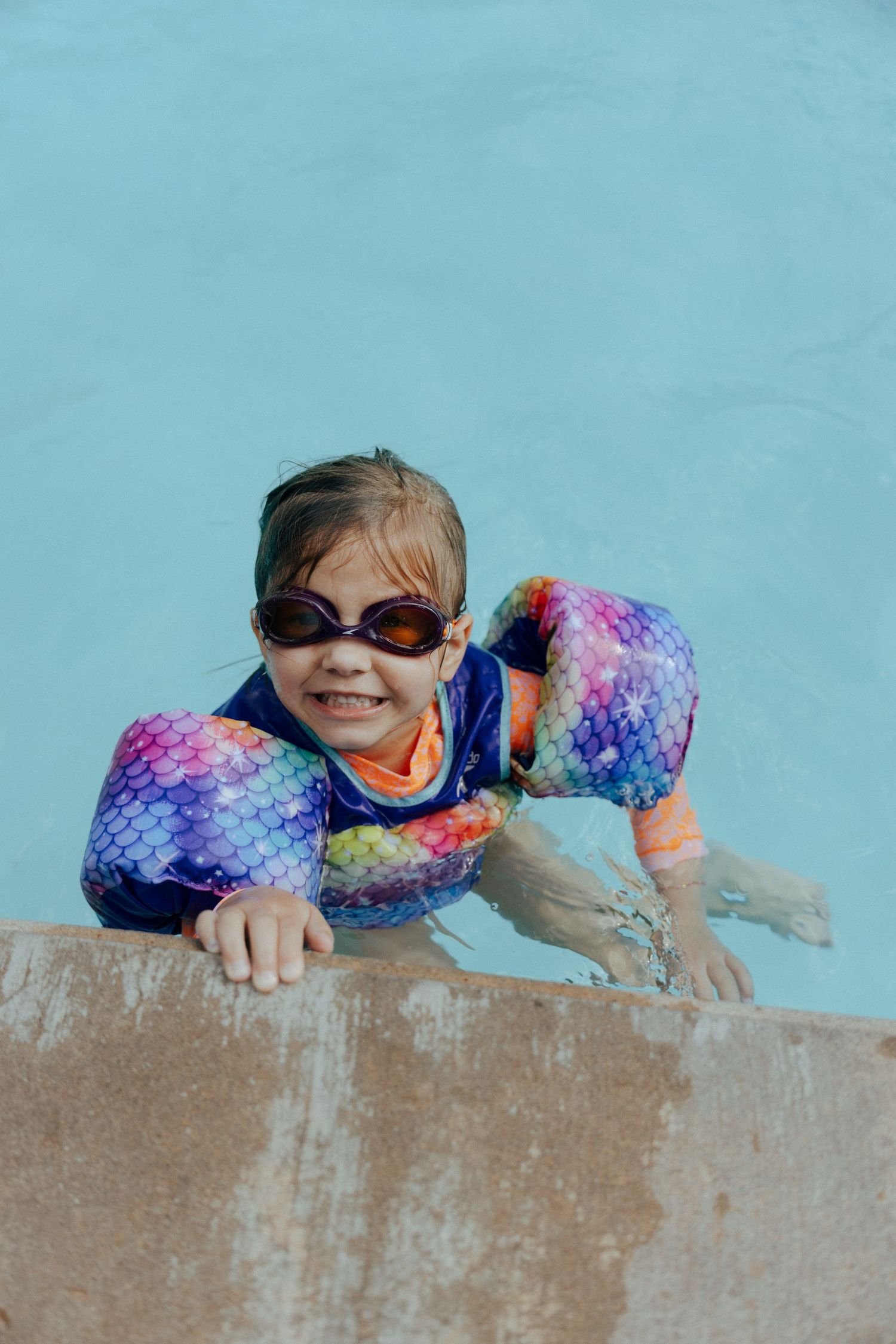 A young swimmer wearing colorful arm floaties and sunglasses hangs on the edge of a pool against a turquoise background.