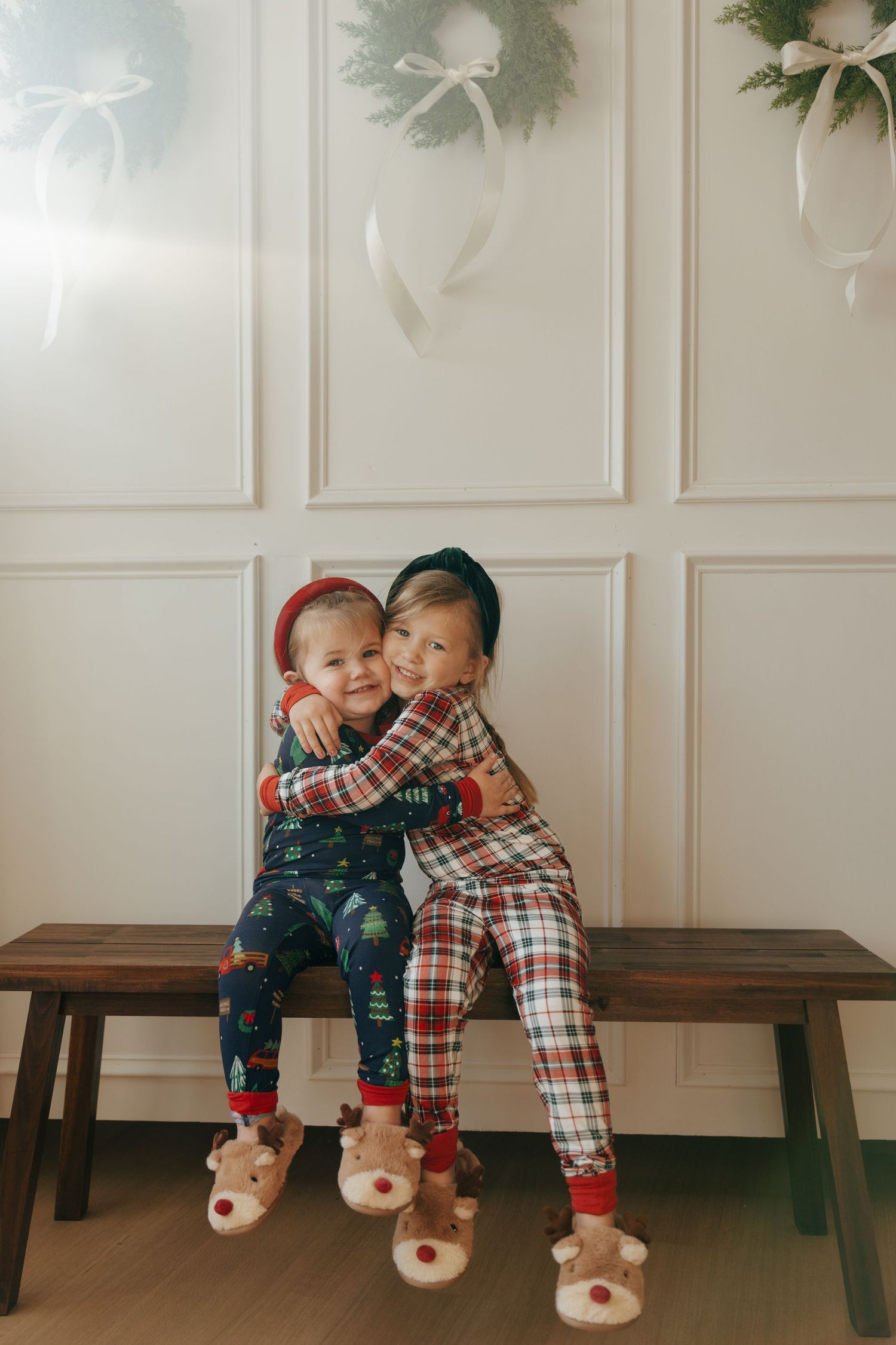 Two children in holiday pajamas and reindeer slippers share a sweet moment while sitting on a wooden bench.