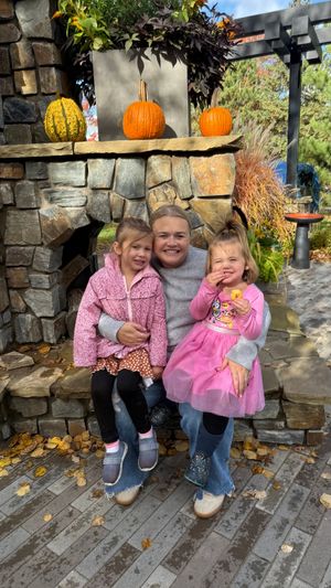 Three children sit together on stone steps decorated with autumn pumpkins and fallen leaves.