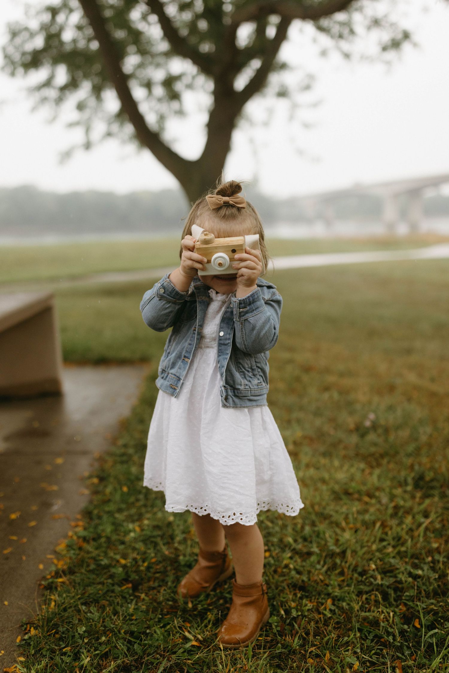 A child in a white dress and brown boots stands in a grassy field while taking photos with a vintage camera.