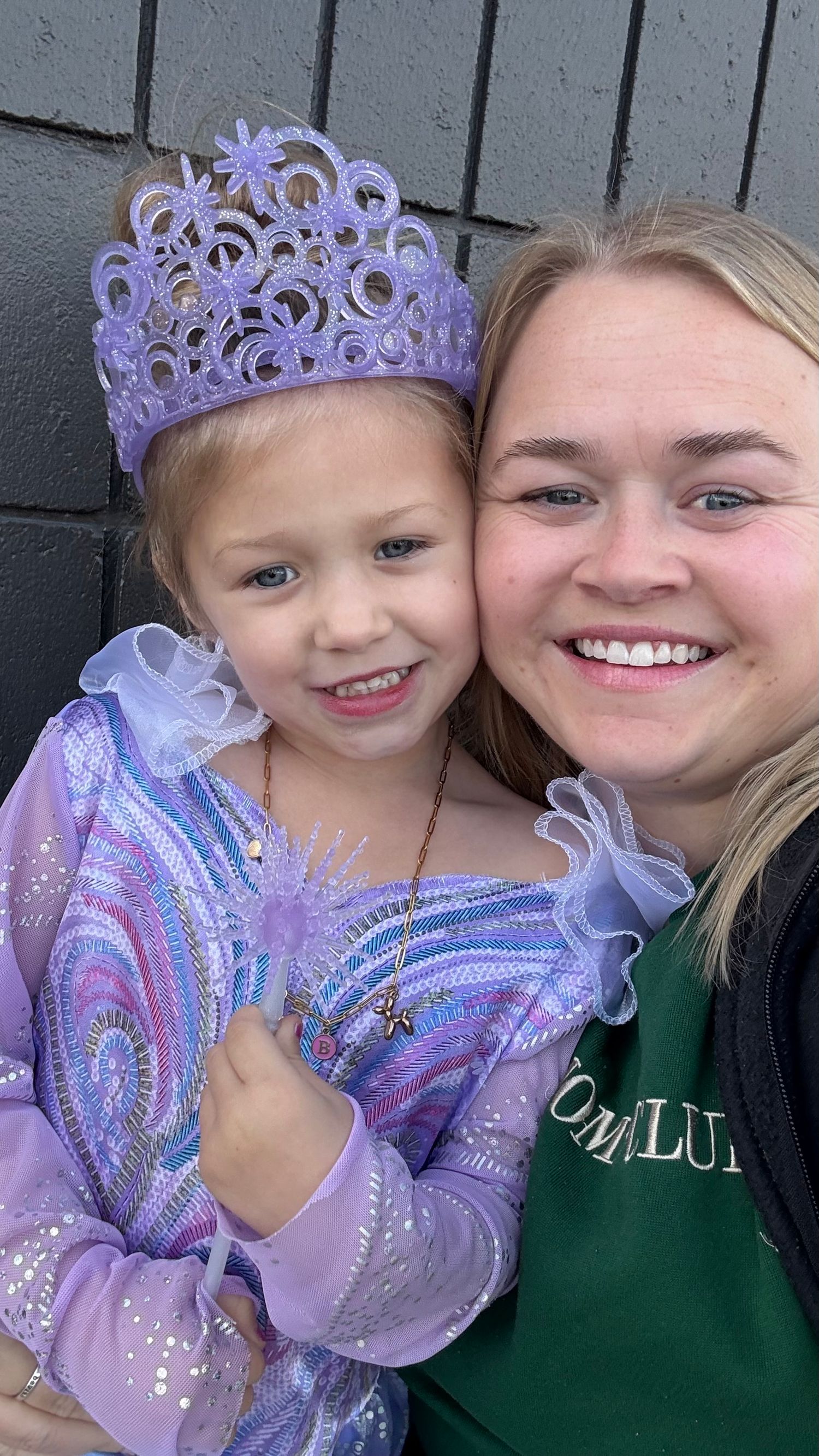 A child wearing a purple princess dress and tiara poses with someone in a green sweatshirt for a selfie photo.