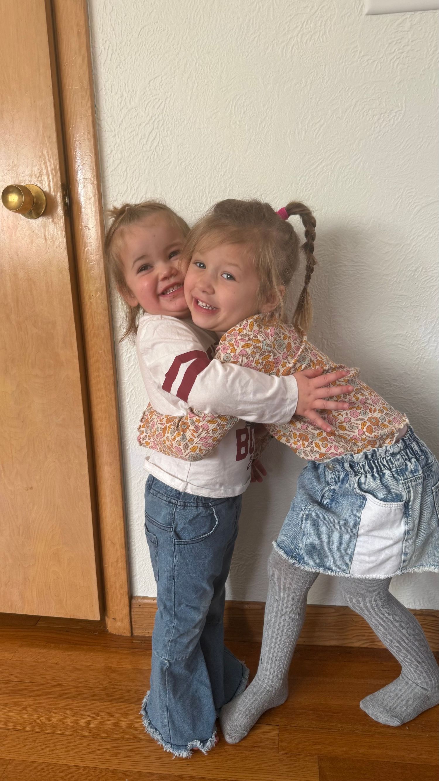Two toddlers in jeans and sweaters playfully hug and laugh against a white wall near wooden cabinets.