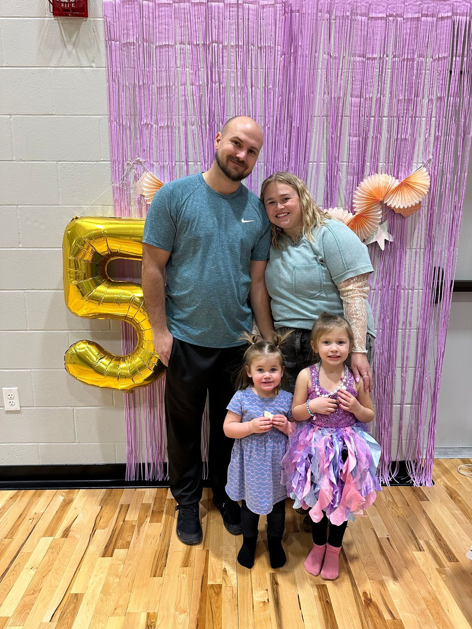 A group poses in front of a pink metallic fringe curtain backdrop with gold number 5 balloon decoration.