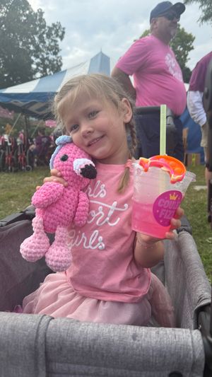 A young child in a pink sweater sits on a gray couch while enjoying a colorful beverage at an outdoor event.