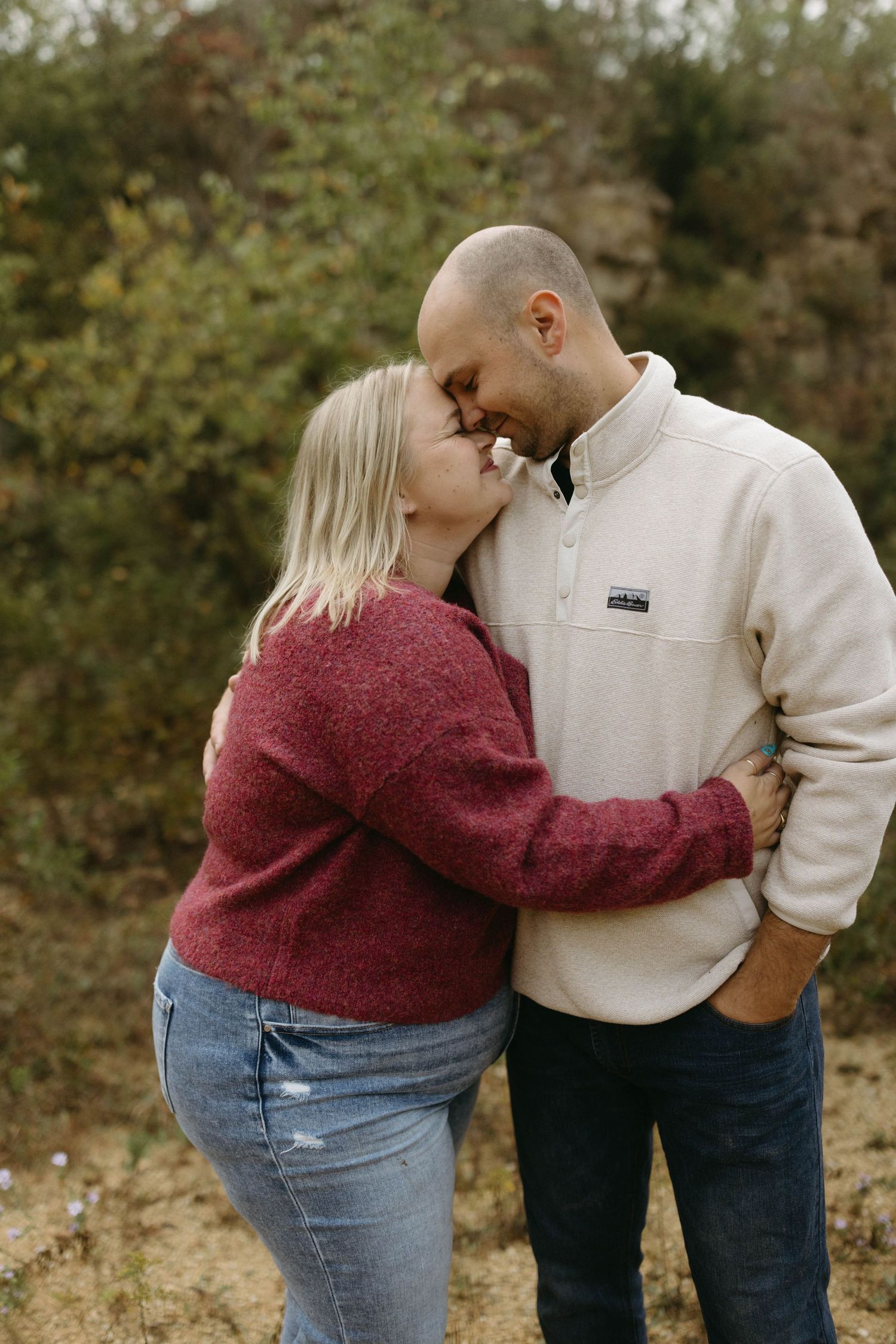 A couple wearing burgundy sweater and cream pullover share intimate moments outdoors against a natural backdrop.