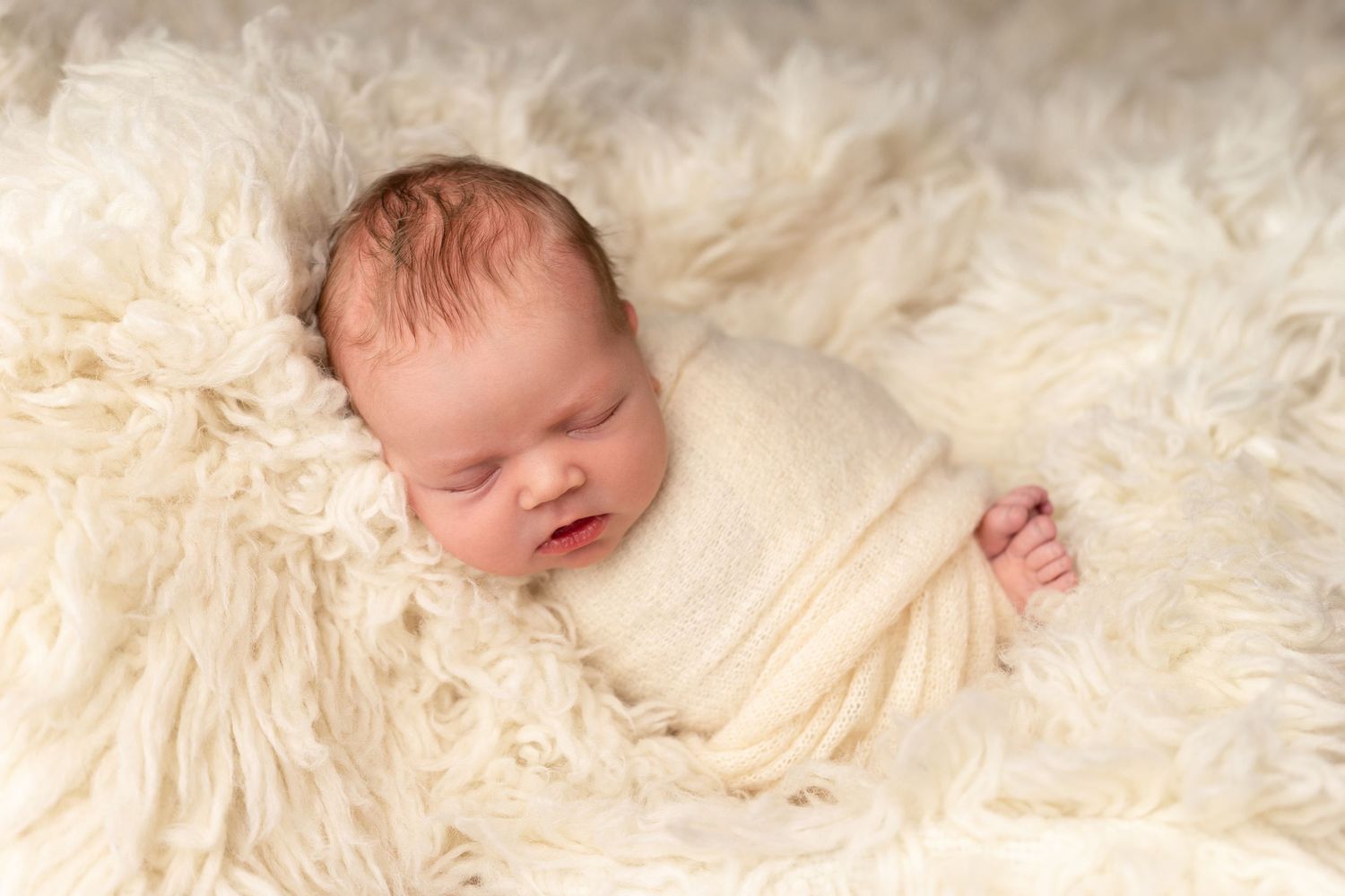 A sleeping newborn baby lies peacefully on soft cream-colored fur blankets in a professional photography setting.