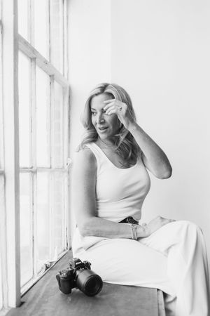 Black and white artistic portrait of someone in white clothing leaning against a bright window ledge.