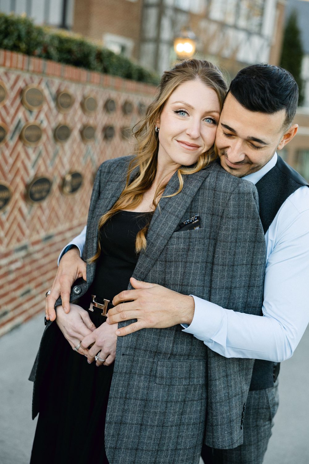Romantic couple embraces while standing against a brick wall in casual elegant attire.