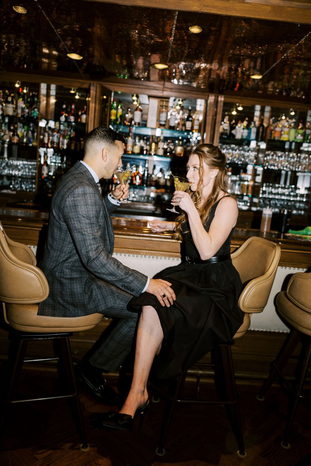 Couple sitting at an elegant bar with leather chairs and shelves of bottles in the background.