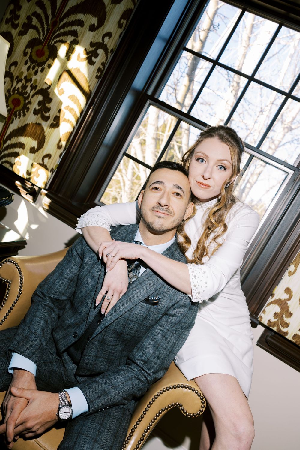 Couple posing together on an elegant couch with ornate wallpaper and window in the background.