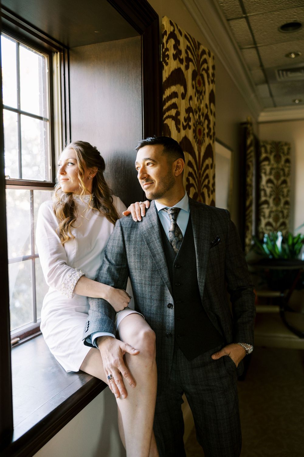 A couple shares a romantic moment by large windows in a vintage-style interior setting.