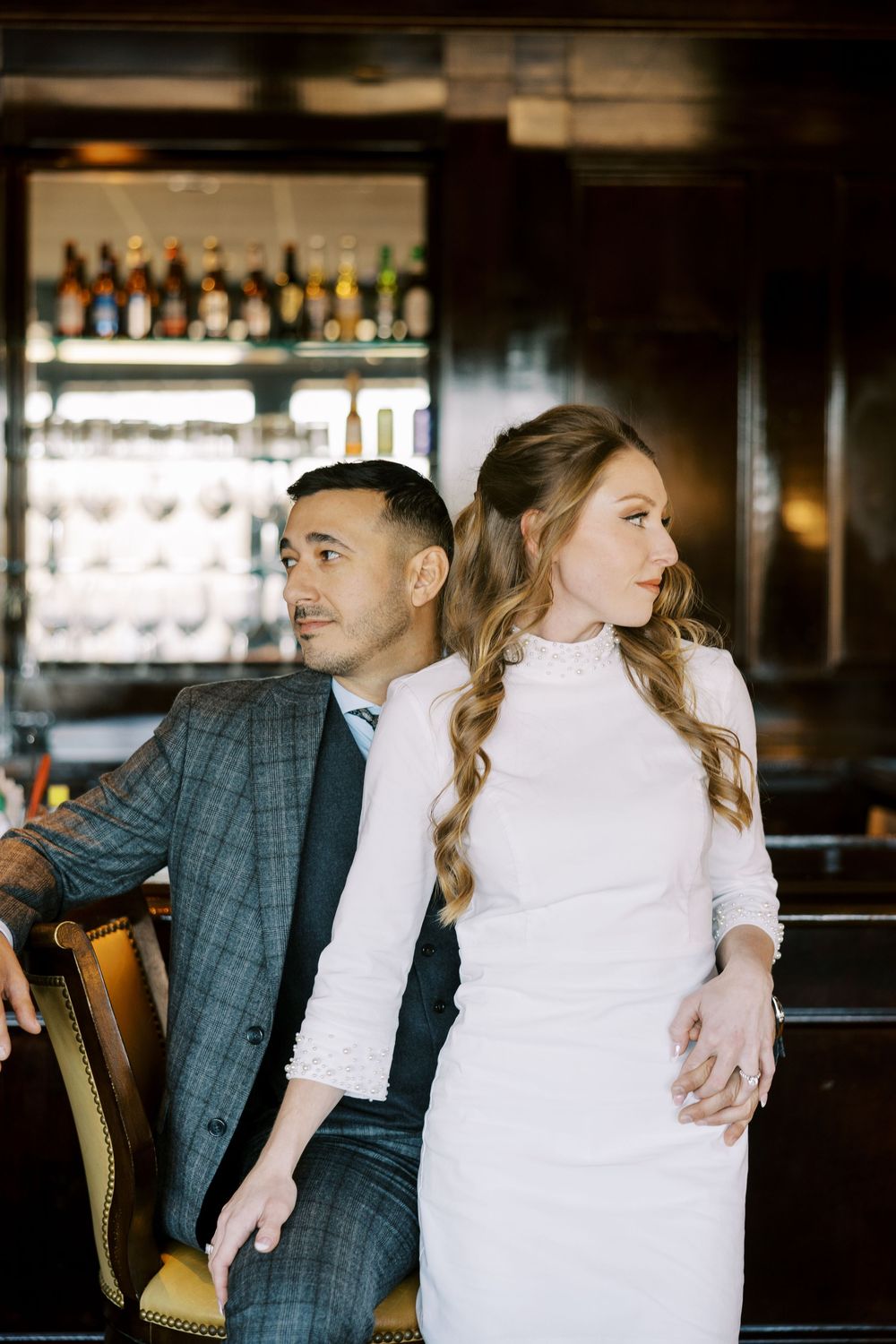 Couple in formal wear poses together at a dimly lit bar with bottles and glassware in the background.