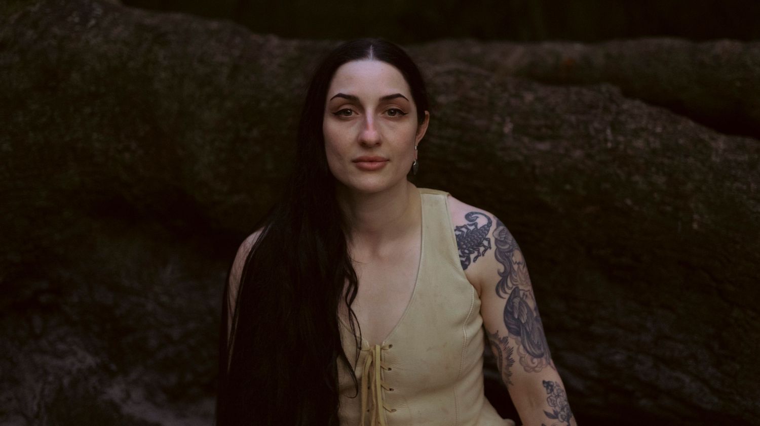 A person in a dark setting with dramatic lighting wearing a beige top and dark clothing.