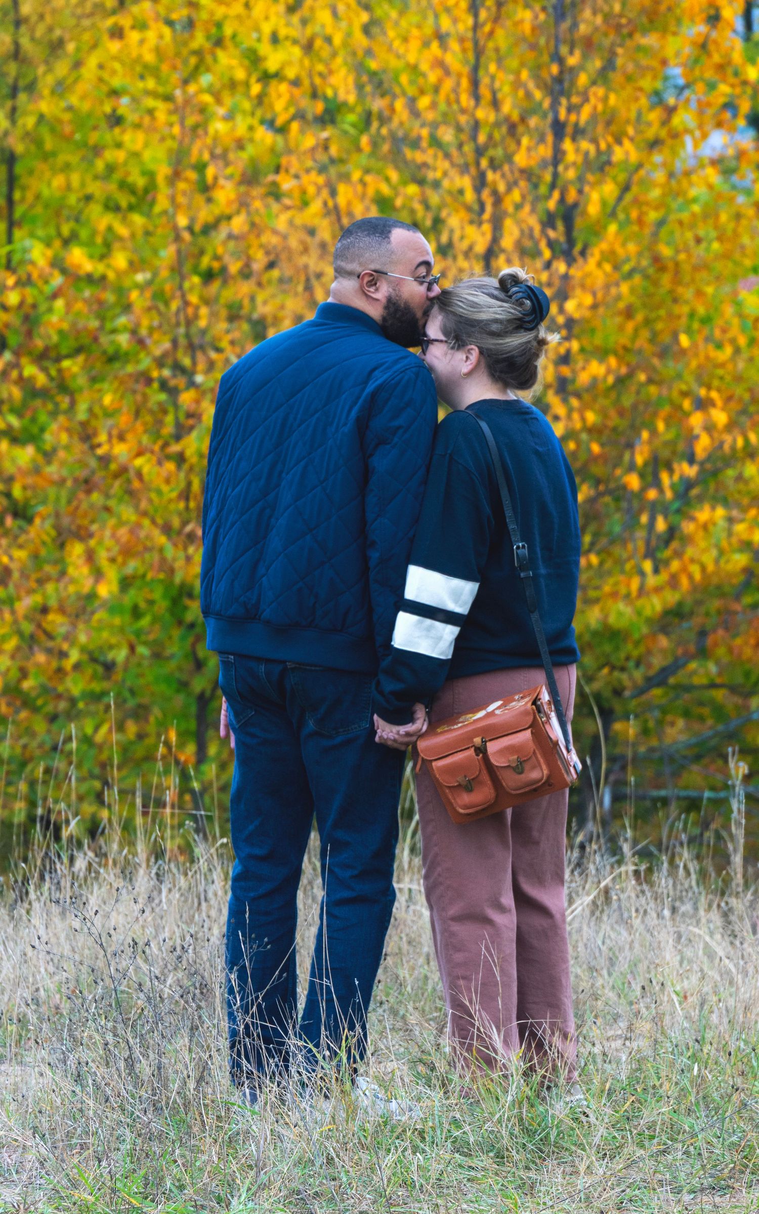 A couple shares an intimate moment in a field with vibrant yellow autumn foliage in the background.