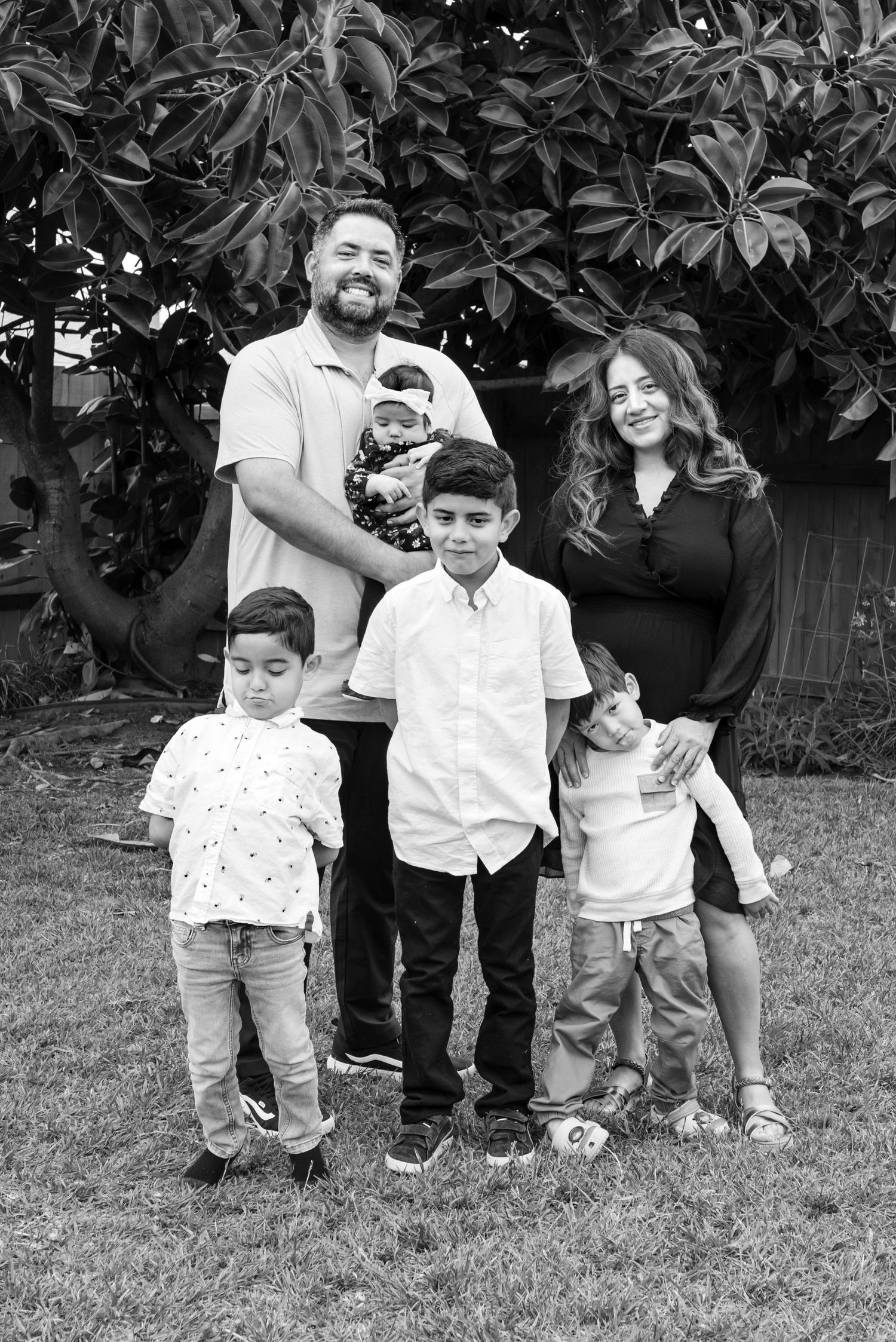 Family standing outside under a huge rubber tree. Dad holds baby while three older boys strike poses in front of the parents. black & white