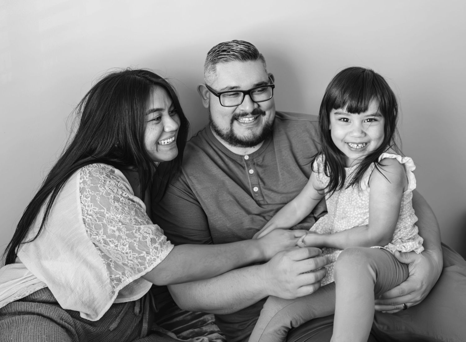 Close up of a smiling family. Toddler sits on dad's lap. Mom lovingly looks at her. (black and white) Doughman Photography