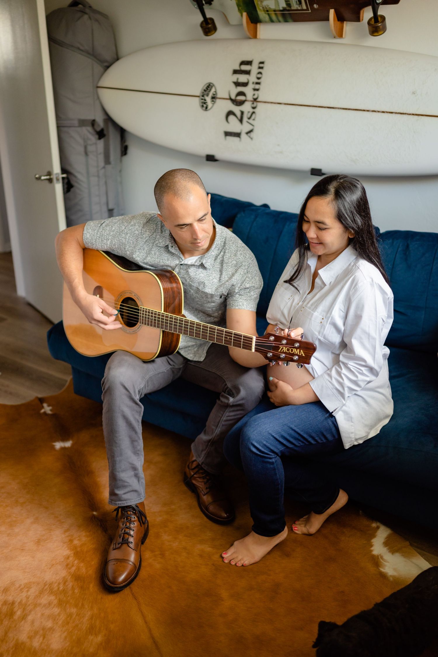 Man sitting on a blue couch with surf board hung on wall, plays guitar to his pregnant wife. In-home maternity session. Doughman Photography