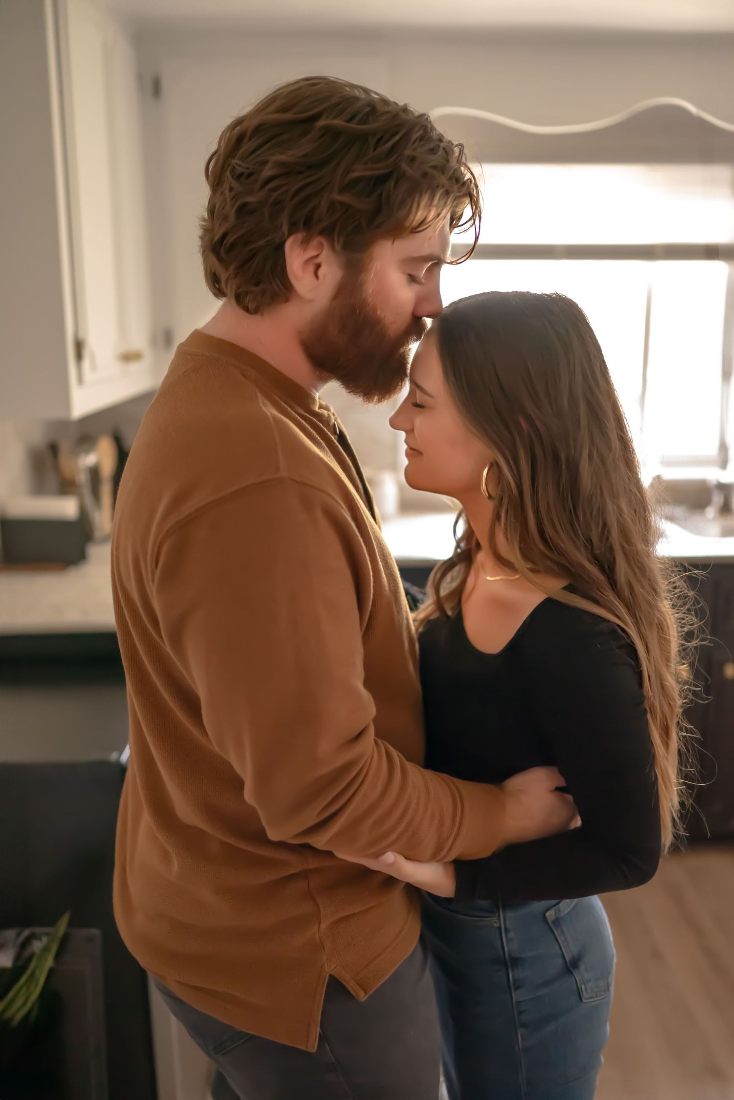 Young couple in the kitchen embrace, he kisses her forehead. In-home couples session.  Doughman Photography