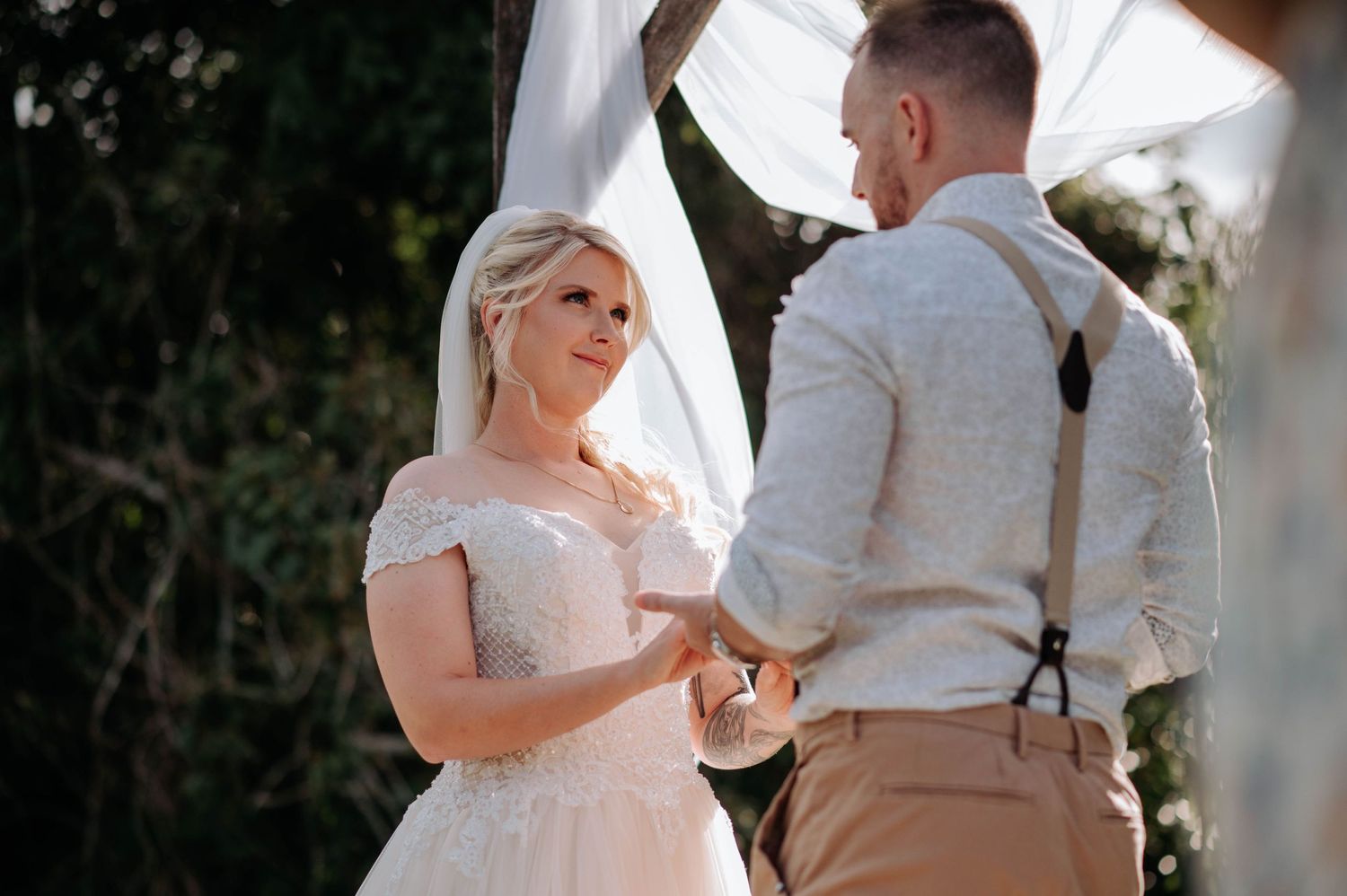 A couple holds hands during their outdoor wedding ceremony under a white draped arch.