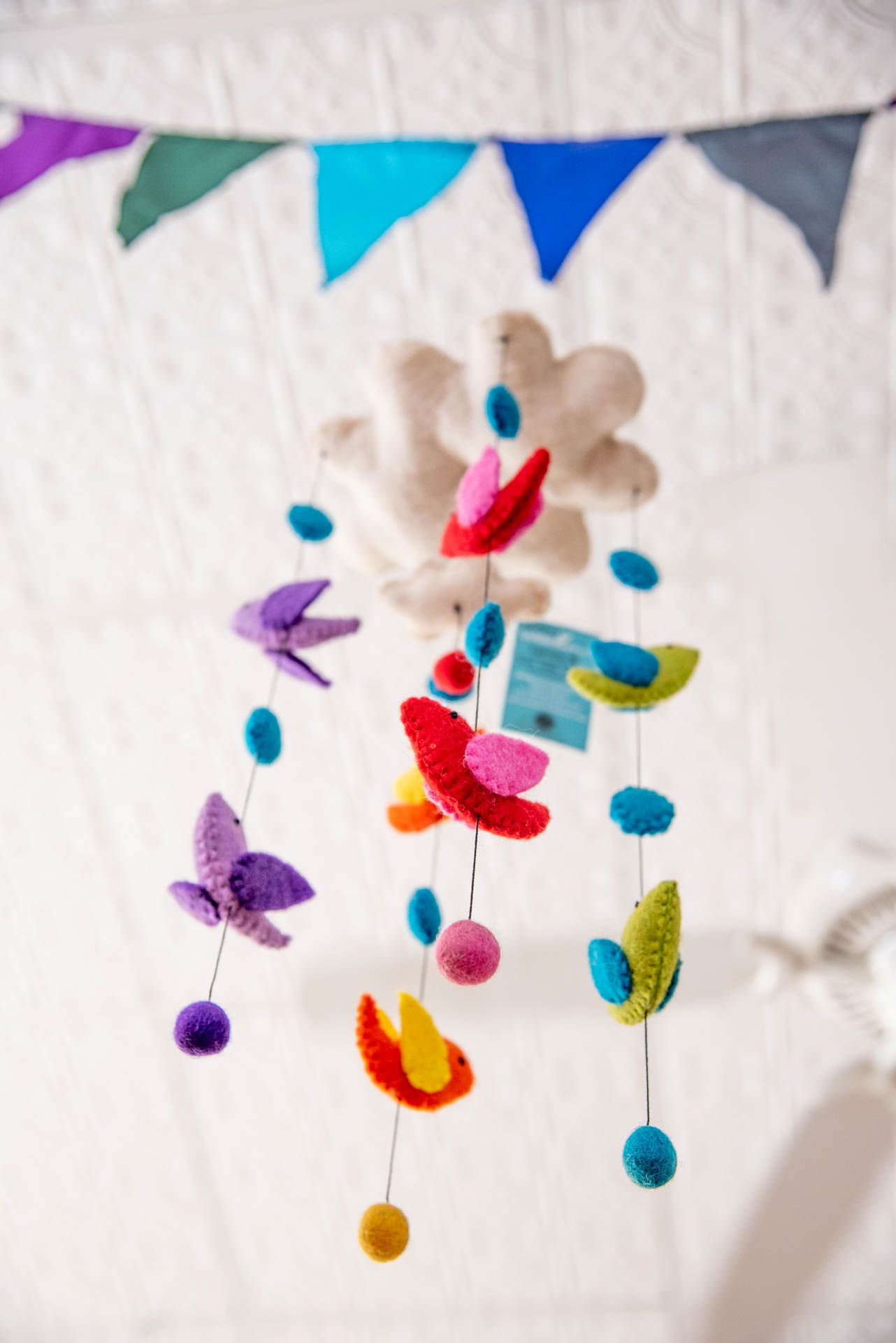 Colorful felt butterfly mobile with hanging pom poms suspended under festive rainbow bunting flags against a white background.
