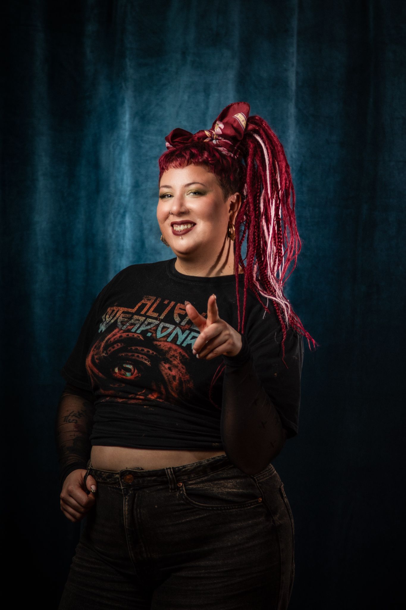 Alternative model in black crop top and jeans poses against blue backdrop with vibrant red dreadlocks and confident smile.