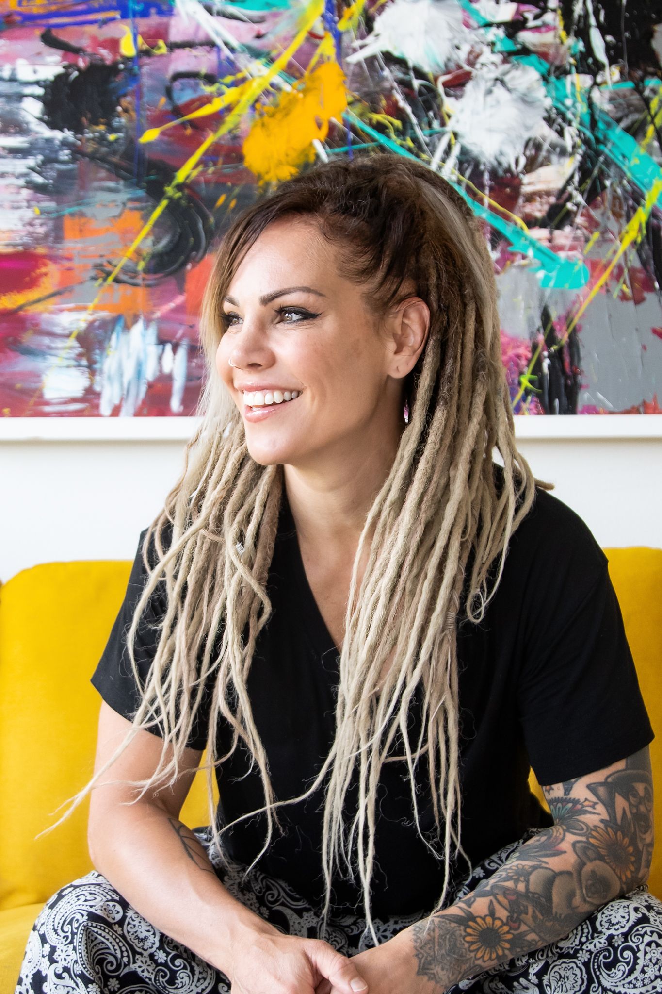 Someone with blonde dreadlocks and tattoos sits on a yellow couch with colorful abstract art displayed on the wall behind them.