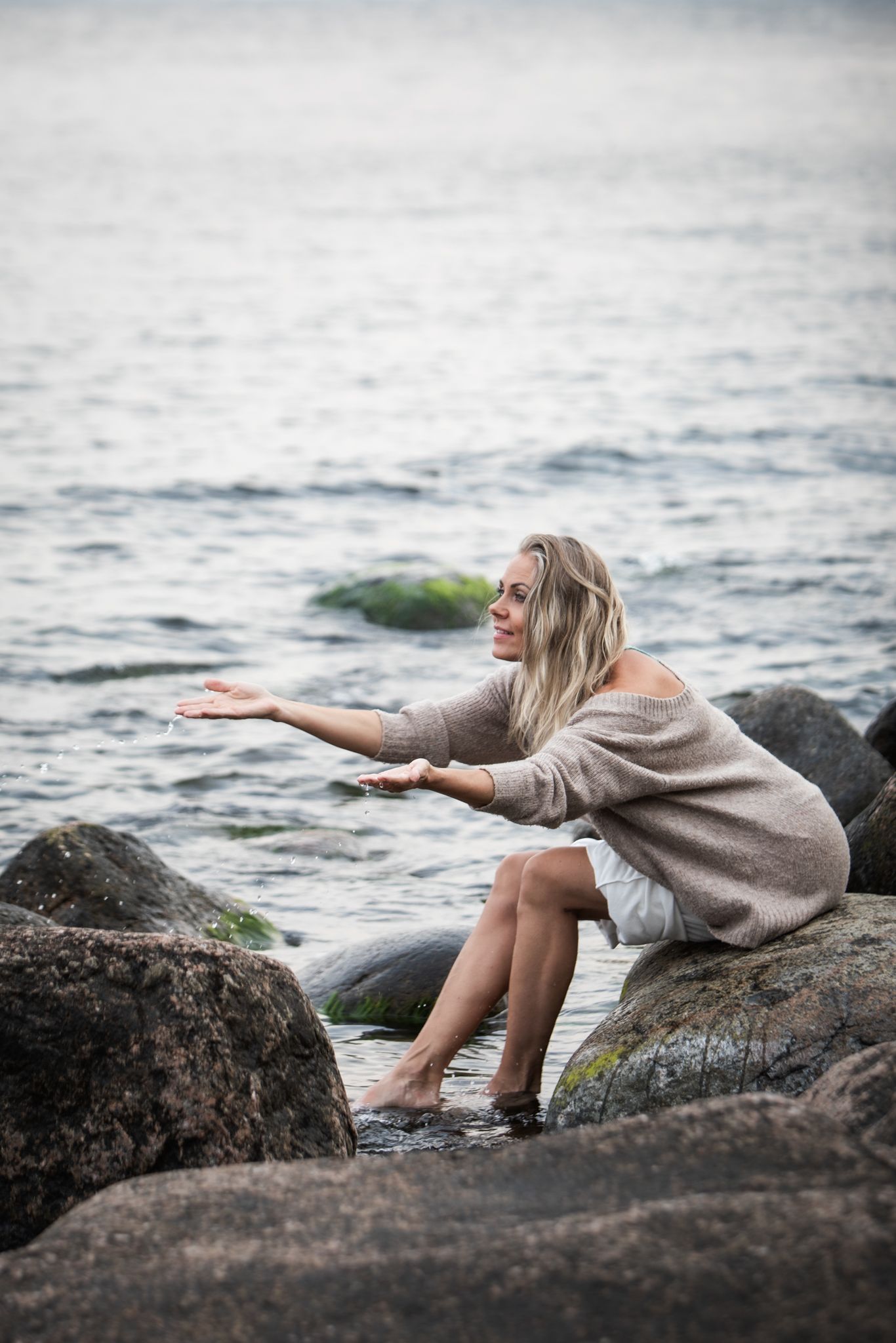 Woman in sweater dress sits on coastal rocks reaching out toward the misty ocean waves.