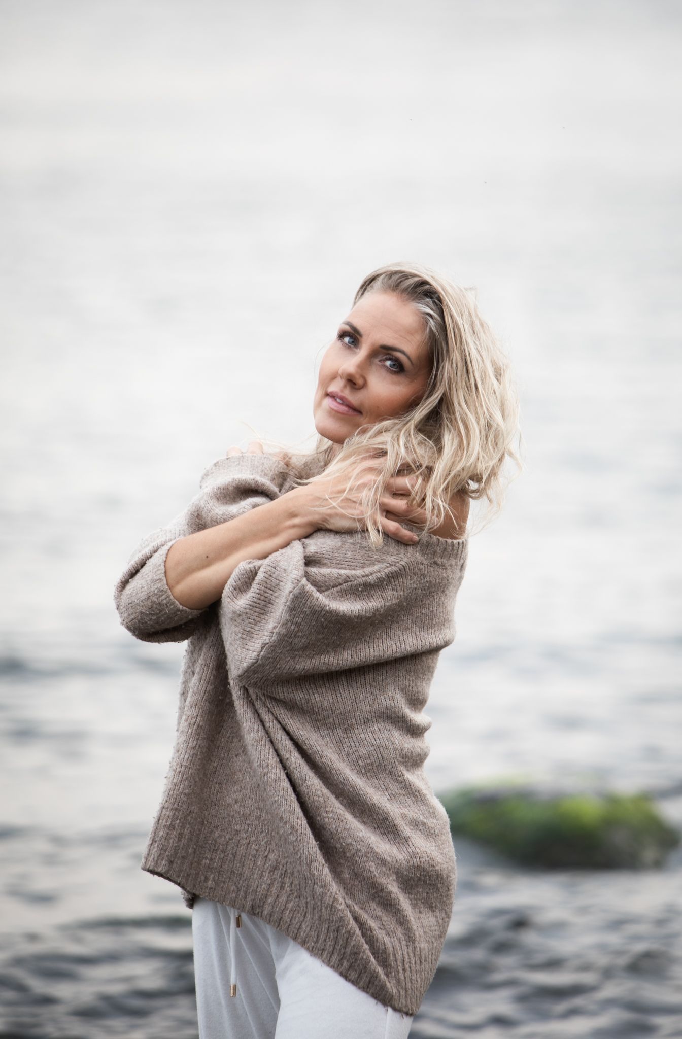 A blonde woman in a cozy beige sweater poses by the ocean on a cloudy day.