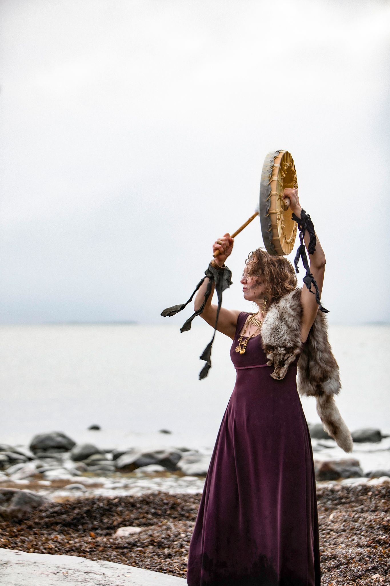 A woman in a burgundy dress holds up a ceremonial drum on a rocky beach while wearing a fur stole.