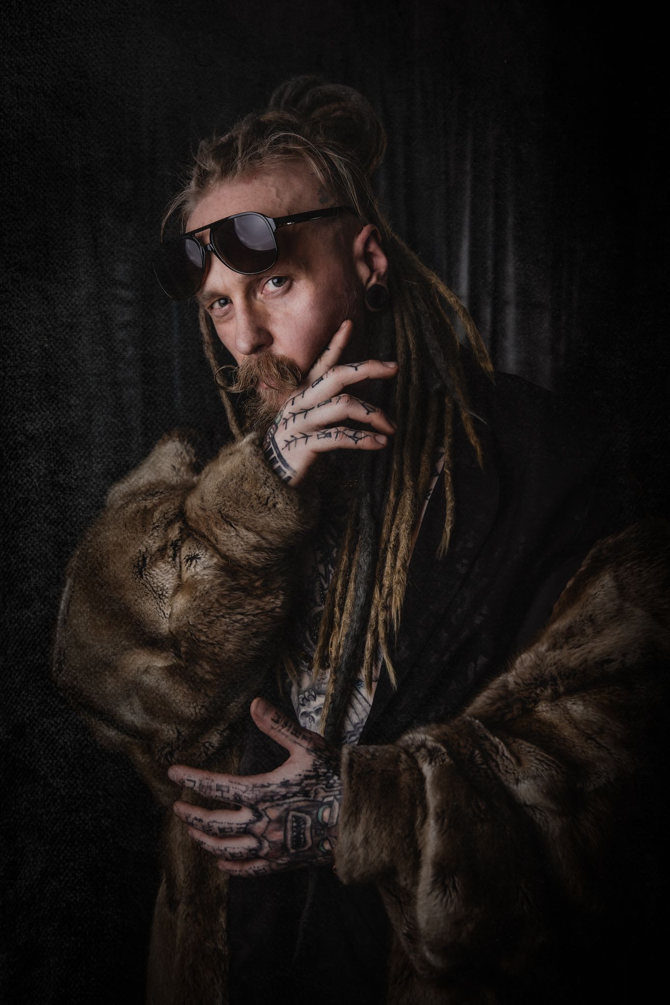 Dramatic portrait in dark tones featuring dreadlocks and fur clothing against a shadowy background.