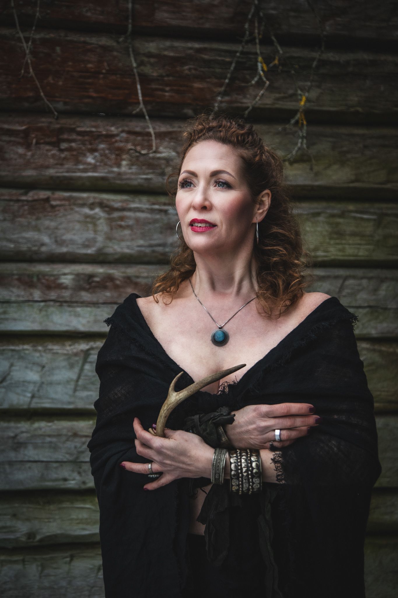 A dramatic portrait of a figure wearing a black shawl with silver bangles and a pendant against a rustic wooden wall.