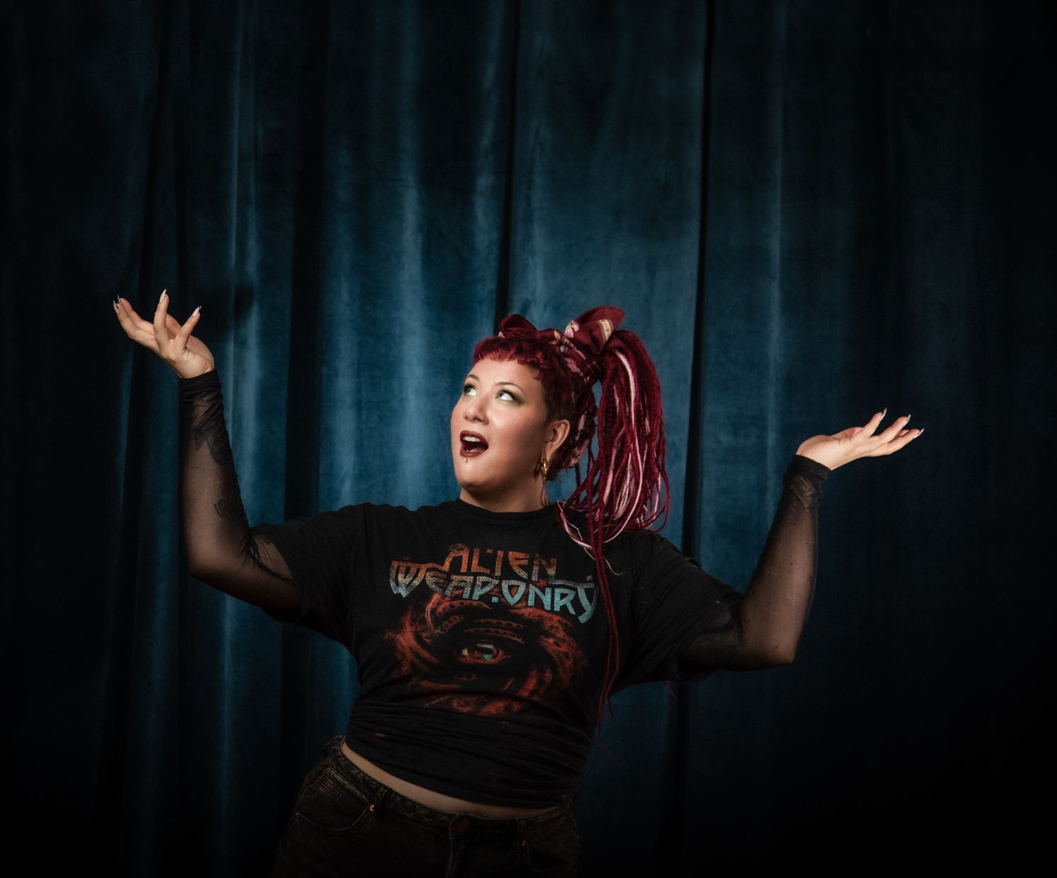 Energetic performer in dark t-shirt with mesh sleeves raises arms dramatically against blue velvet curtain backdrop.
