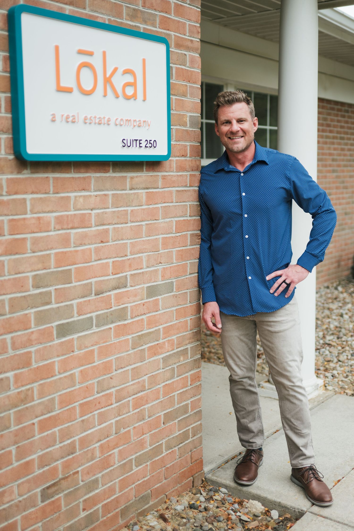 A person in a blue button-down shirt and khakis stands next to a brick wall with a sign for 'Lokal' business suite.