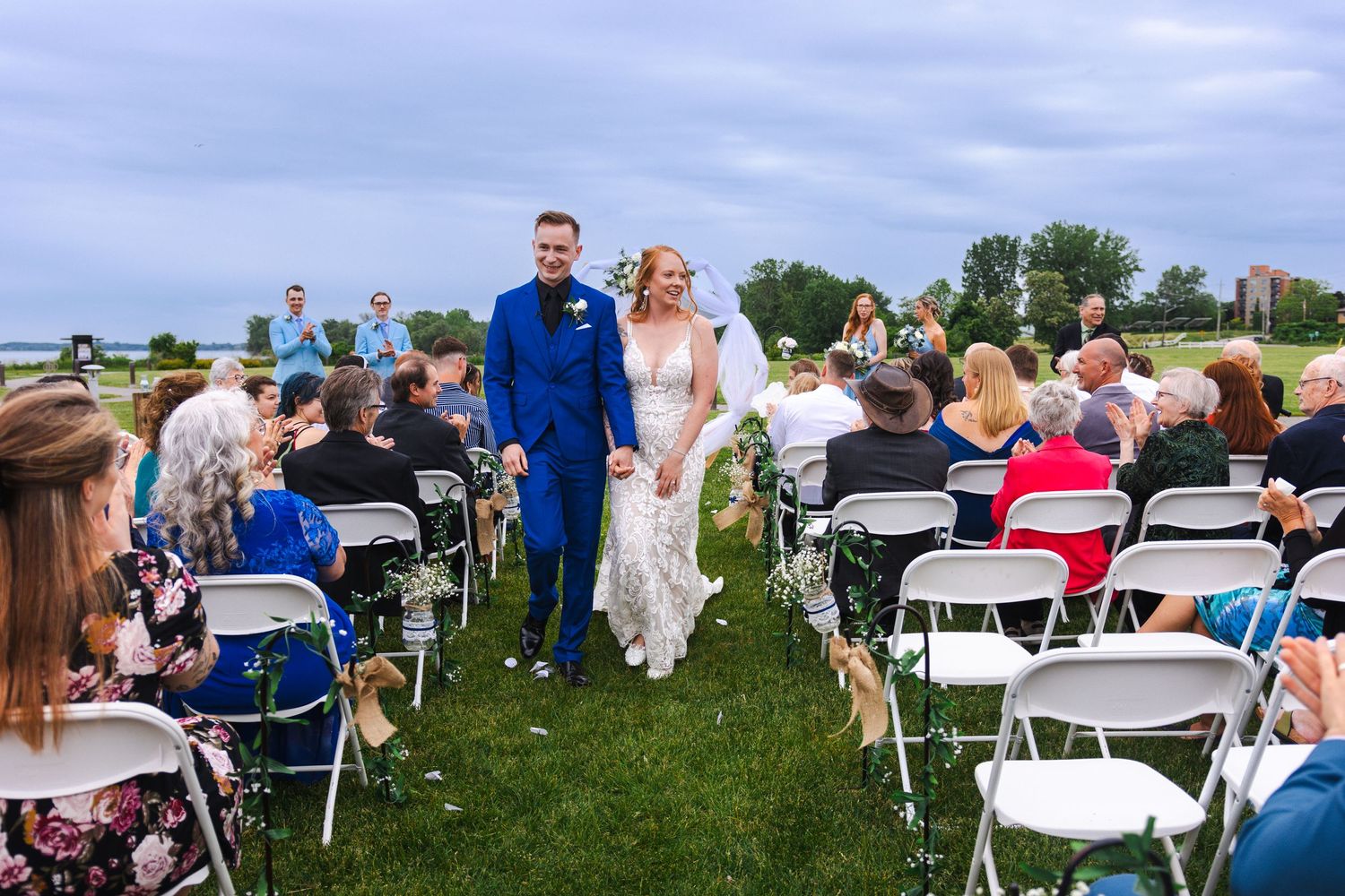 A couple walks down the outdoor wedding aisle between white chairs as guests look on under a blue summer sky.