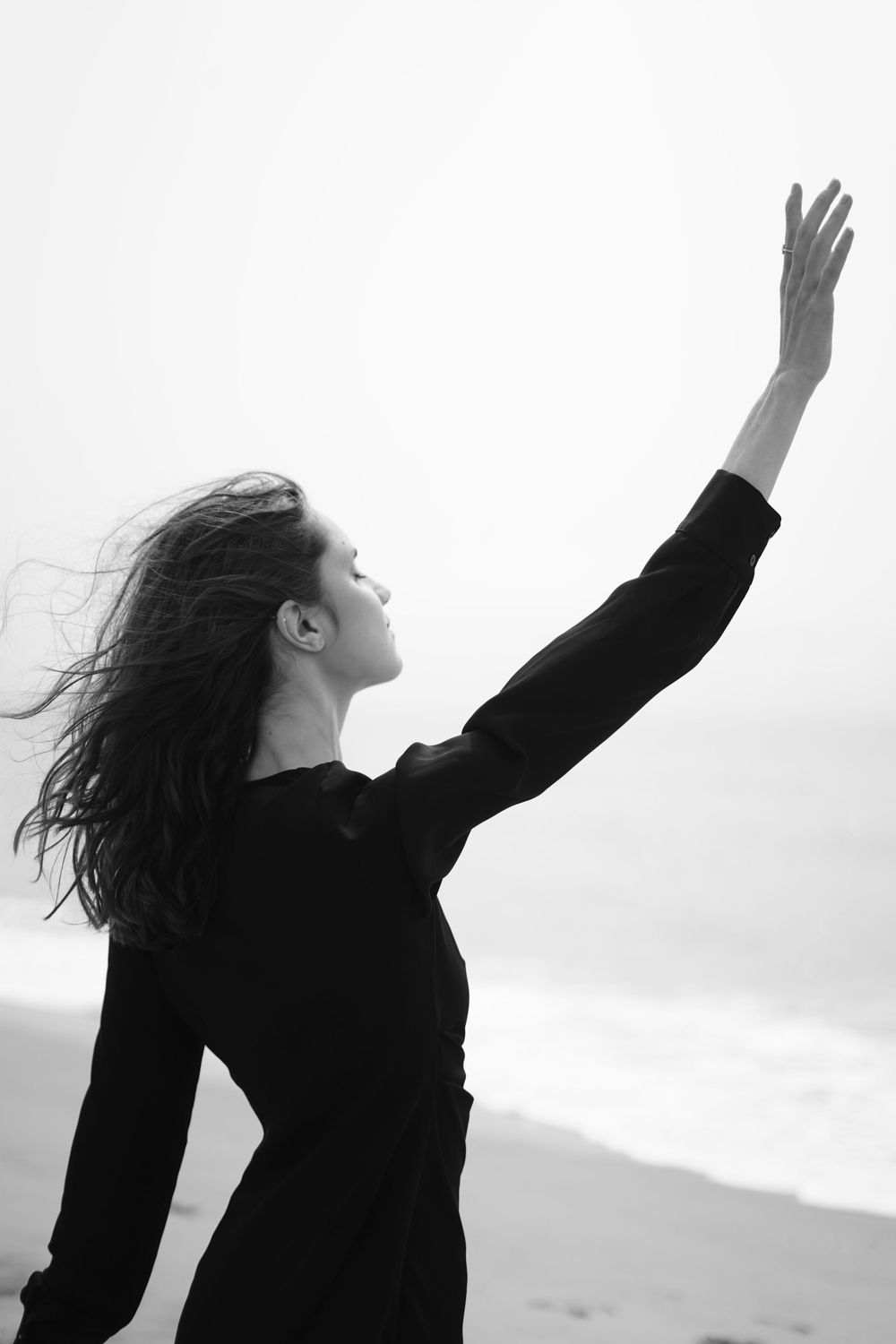 A silhouette of a figure with flowing hair reaching up against a bright sky in black and white.