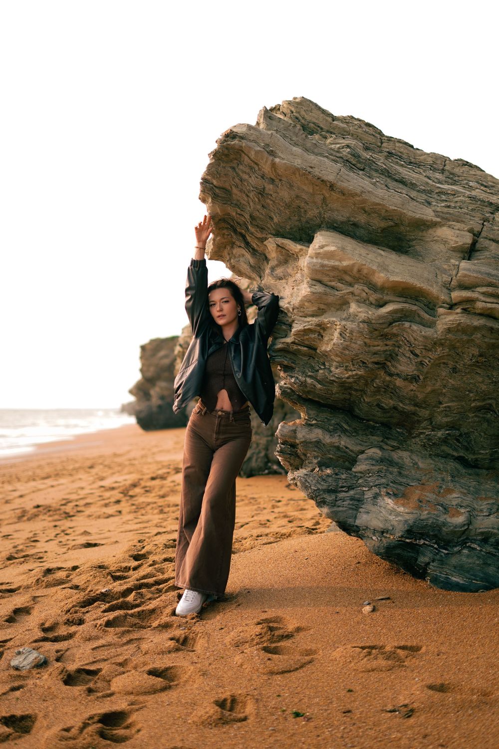 A person reaches upward while posing next to a large rock formation on a beach at sunset.