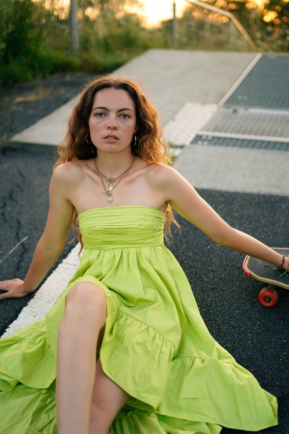 Person in bright lime dress posing with skateboard on asphalt at sunset.