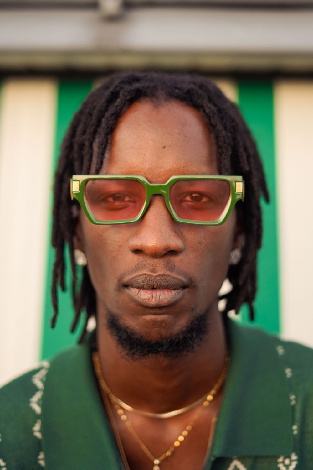 Portrait featuring bright green sunglasses and dreadlocks against striped background.