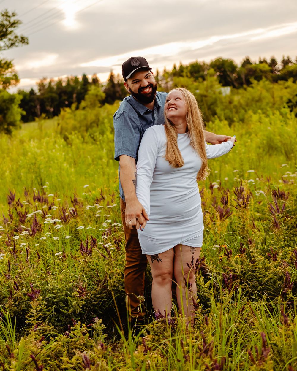 A couple shares a joyful moment together while standing in a wildflower meadow at sunset with forest in background.