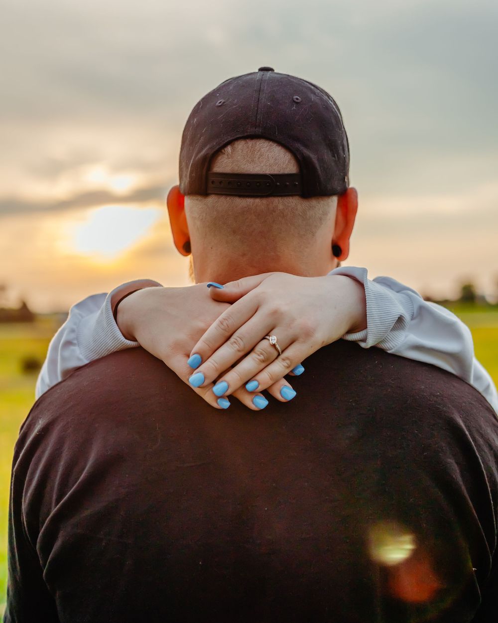 Person wearing a baseball cap embraces someone from behind while watching a golden sunset in a grassy field.