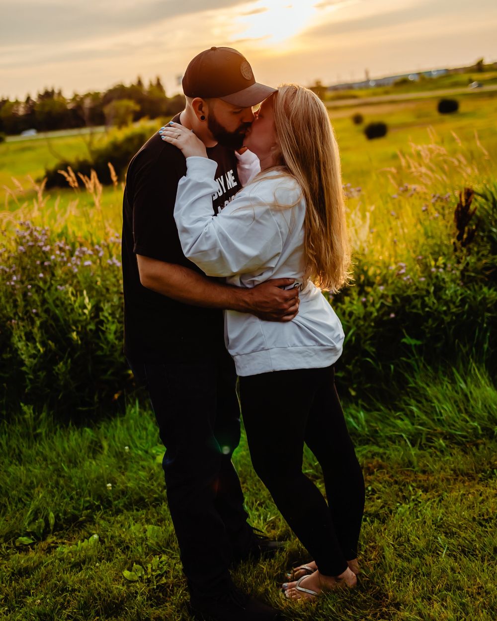 Couple embraces in golden meadow during sunset while surrounded by tall grass and hay bales.