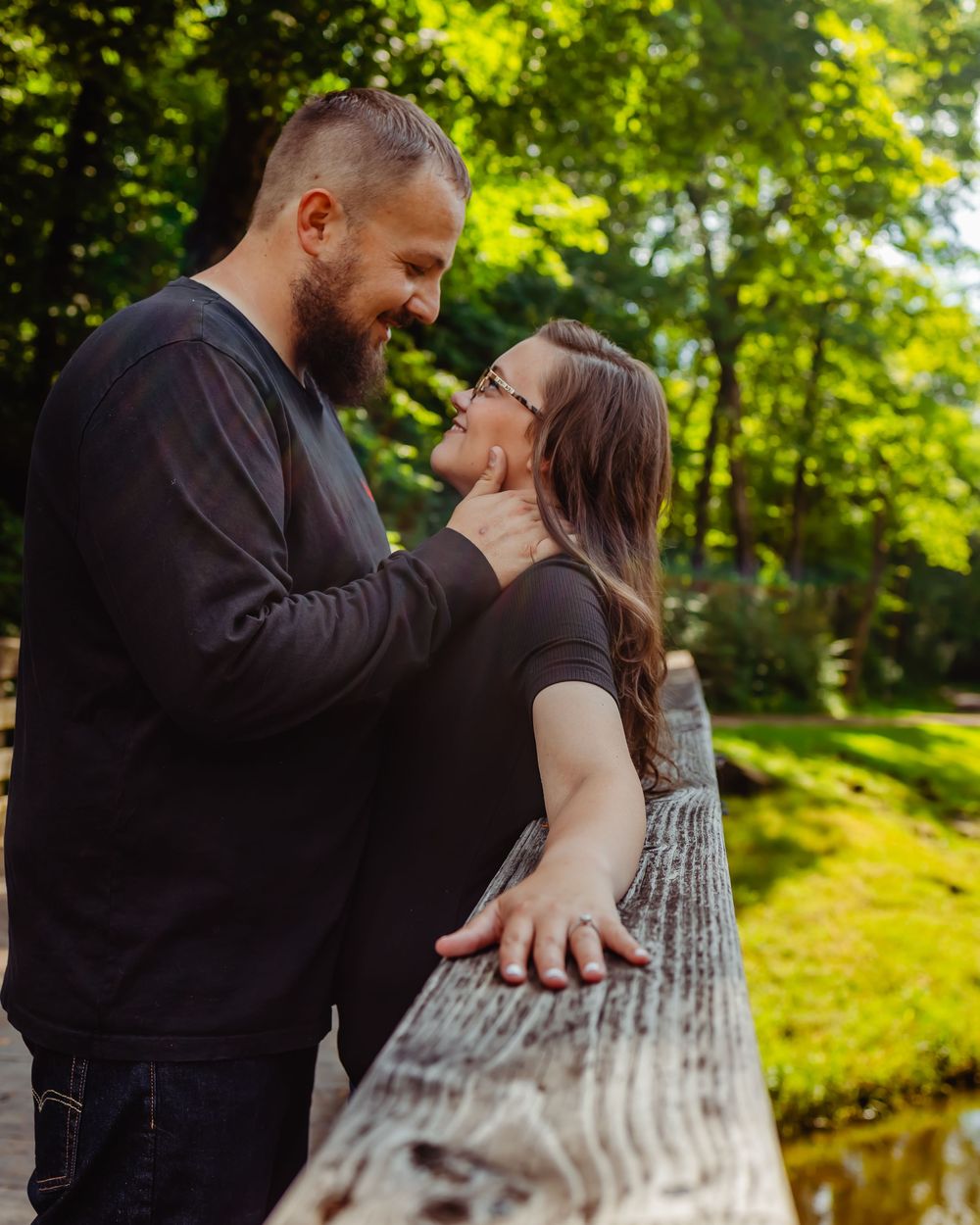 A couple shares a romantic moment leaning against a wooden railing in a sunlit forest setting.