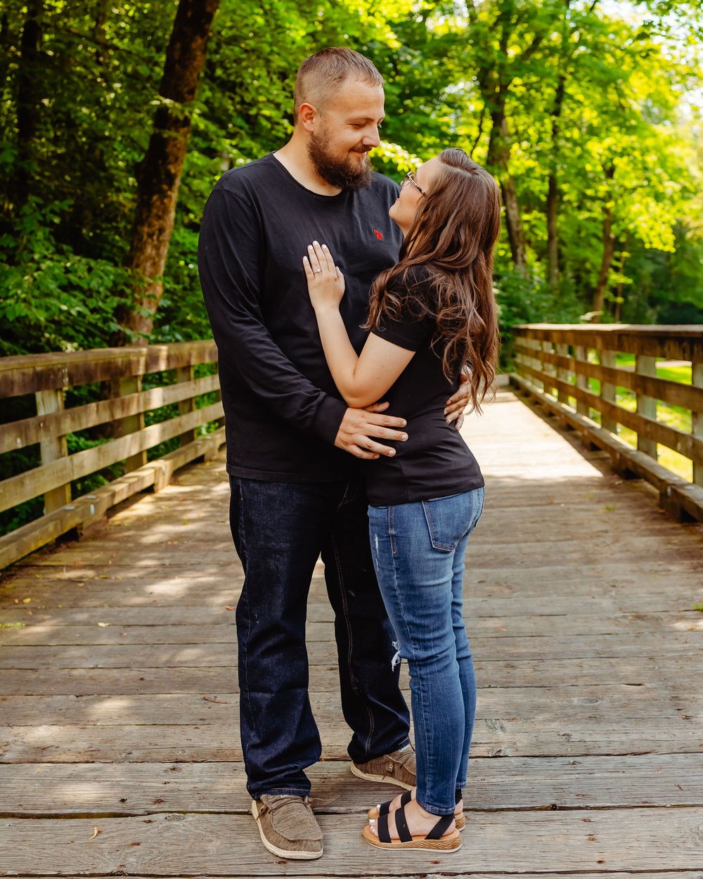 A couple in matching black tops share an intimate moment on a wooden bridge surrounded by lush green forest.