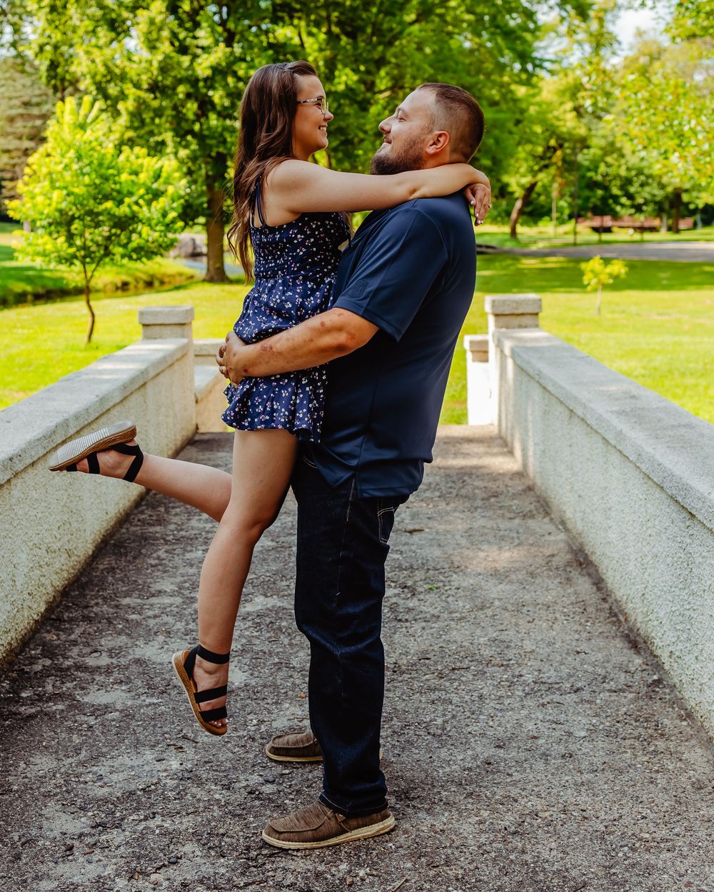 A romantic couple embraces on a stone pathway surrounded by lush green trees and foliage in a park setting.