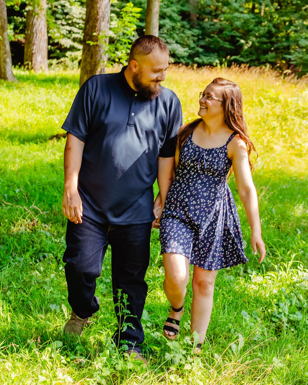 Two people walk together on a grassy path in a sunny wooded area, wearing casual summer clothing and smiling at each other.